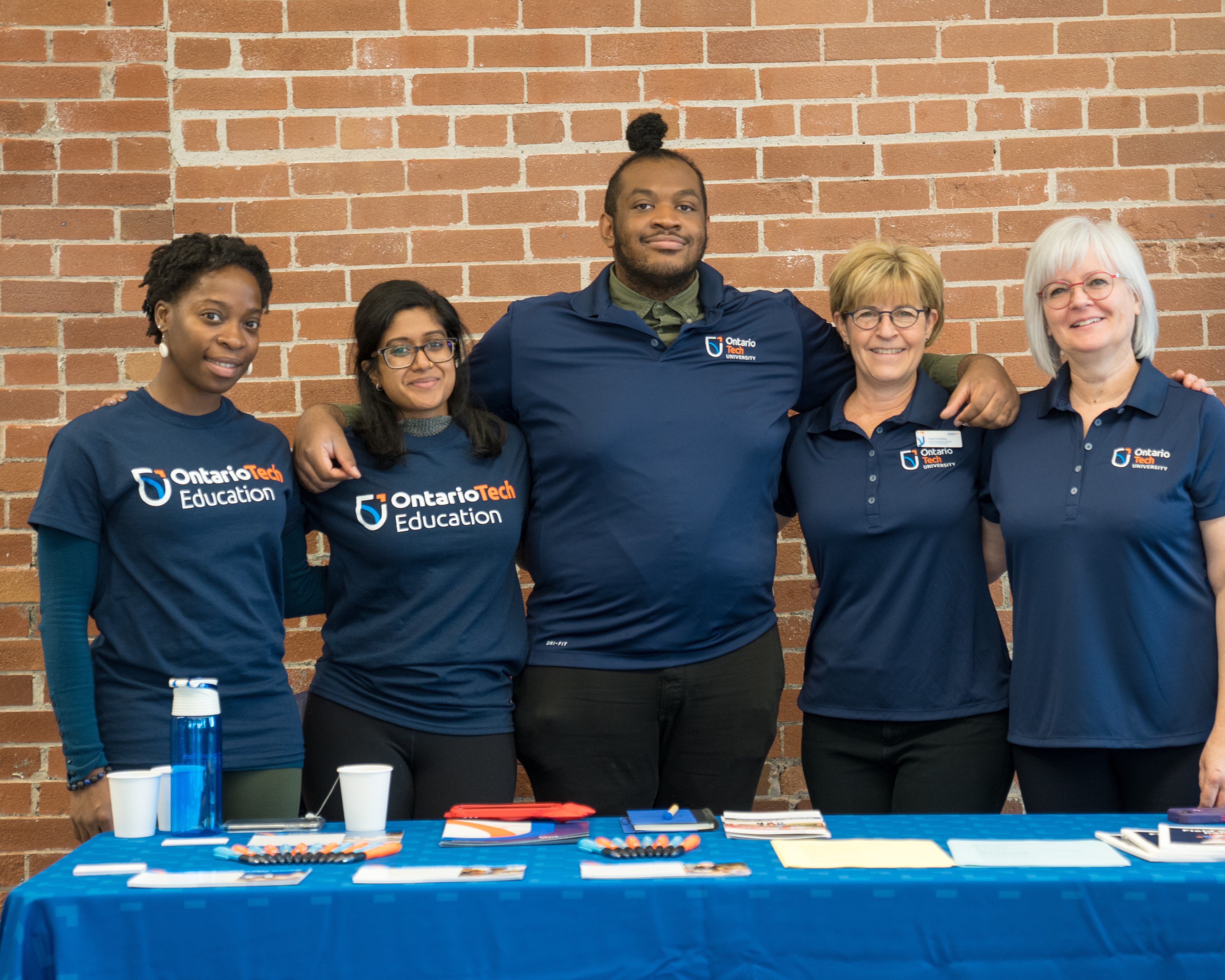 Four smiling individuals wearing matching navy blue shirts pose together behind an information table. The table is covered with pamphlets and promotional items, set against a brick wall background. 